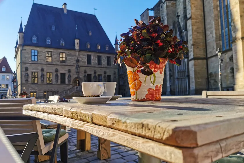 Espresso und ein bunter Blumentopf auf einem sonnigen Terrassentisch, im Hintergrund das Osnabrücker Rathaus